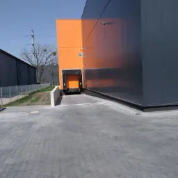 Zewnętrzny widok na rampę załadunkową pomarańczowo-szarego magazynu z kostki brukowej, widoczne drzewa bez liści w tle.
