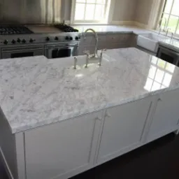 Wyspa kuchenna z białym marmurowym blatem, zlewem i baterią w nowoczesnej kuchni z szarymi szafkami i dużym piecem.