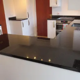 Czarny, błyszczący blat kuchenny z drobnymi, jasnymi inkluzjami, zamontowany w białej zabudowie kuchennej, z widocznym zlewem i fragmentem płyty grzewczej.