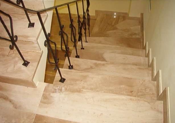 Wewnętrzne schody z beżowego marmuru z kutą, czarną balustradą, widok z góry na dół.