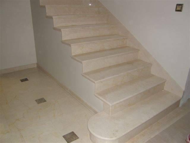 Beżowe, marmurowe stopnie schodów wewnętrznych z zaokrąglonym dolnym stopniem, dopasowane kolorystycznie do podłogi z kwadratowymi wstawkami, oświetlenie dzienne.