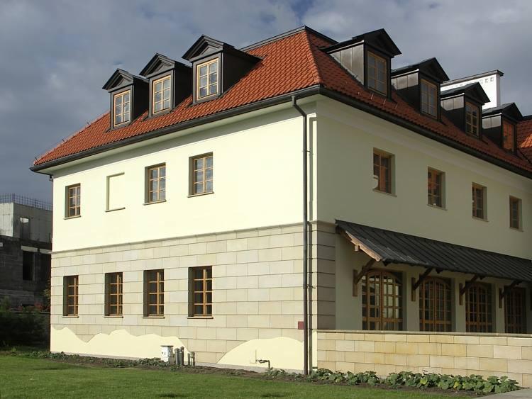 Elewacja budynku z jasnego kamienia z czerwonym dachem i oknami w stylu dworkowym, widoczny fragment trawnika i budowa w tle.