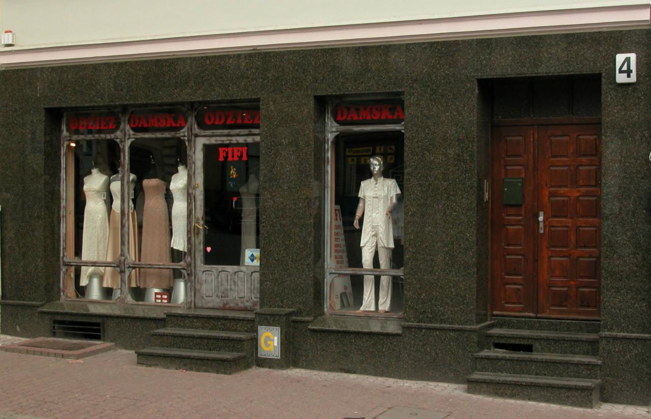 Fasada sklepu odzieżowego z kamienną okładziną, wystawa z trzema manekinami w sukniach wieczorowych, szyld z napisem 'ODZIEŻ DAMSKA' i brązowe drewniane drzwi wejściowe z numerem 4.