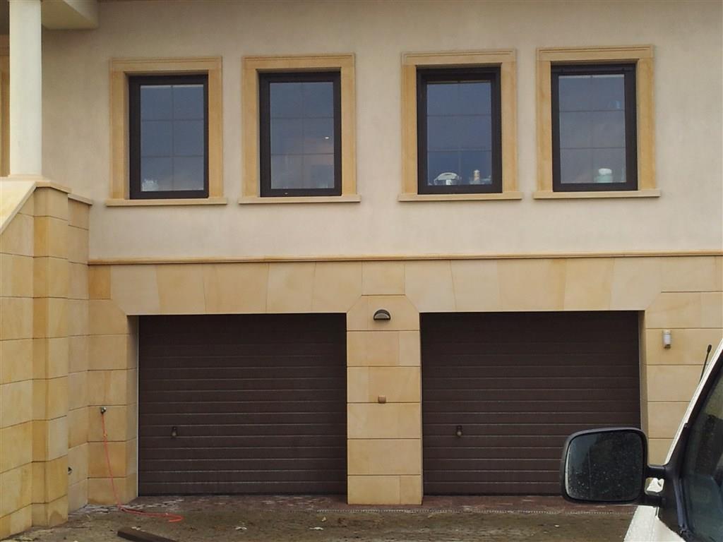 Elewacja budynku z piaskowca z czterema oknami i dwoma bramami garażowymi, widoczne detale wykończeniowe z kamienia.