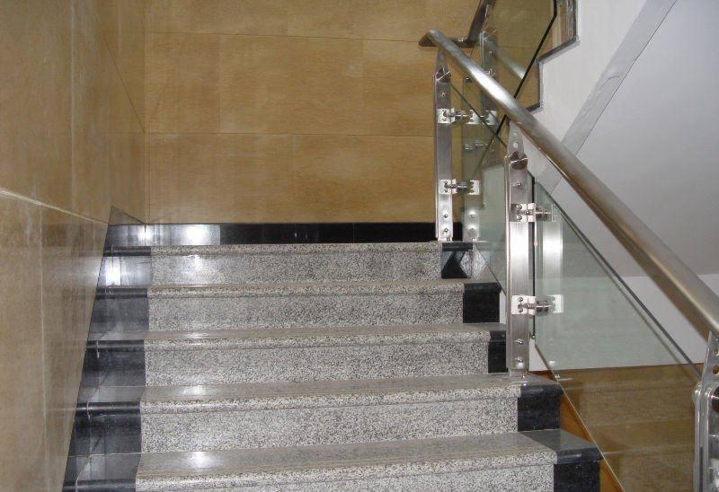 Granitowe schody z czarnymi podstopnicami i szklaną balustradą ze stalowymi mocowaniami, oświetlone światłem dziennym.