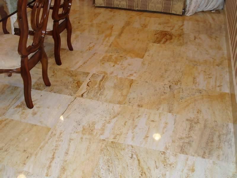 Podłoga wyłożona jasnymi, beżowo-złotymi płytami kamiennymi, widoczne krzesła z ciemnego drewna w tle oraz fragment sofy.