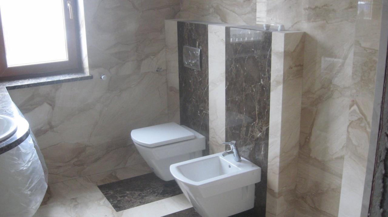 Wykończona łazienka z marmurowymi płytkami, wiszącą toaletą i bidetem, brązowo-białe pasy na ścianie za urządzeniami sanitarnymi, umywalka częściowo zasłonięta folią ochronną.