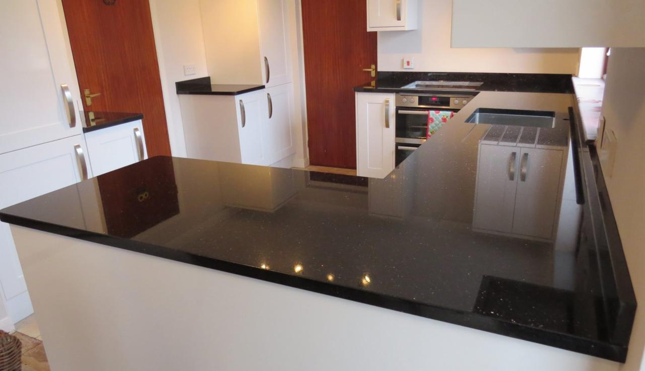 Czarny, błyszczący blat kuchenny z drobnymi, jasnymi inkluzjami, zamontowany w białej zabudowie kuchennej, z widocznym zlewem i fragmentem płyty grzewczej.