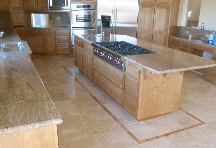 Jasna kuchnia z drewnianą zabudową, wyspą z blatem z kamienia i płytkami podłogowymi w odcieniach beżu, z widoczną kuchenką gazową.