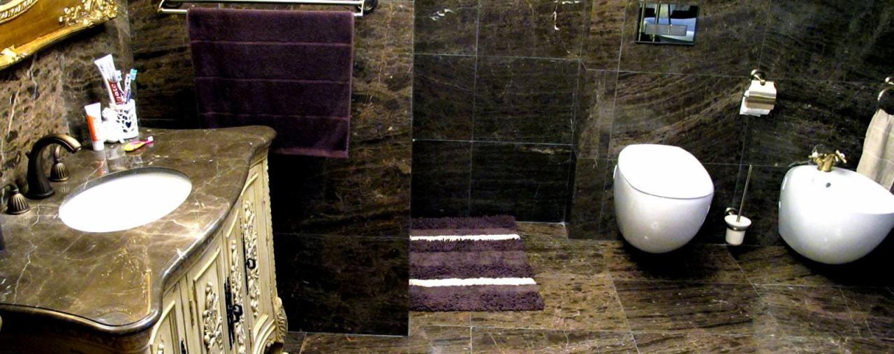 Elegancka łazienka wykończona ciemnym marmurem, z umywalką w ozdobnej szafce, toaletą i bidetem o nowoczesnym kształcie oraz fioletowymi akcentami w postaci ręcznika i dywanika.