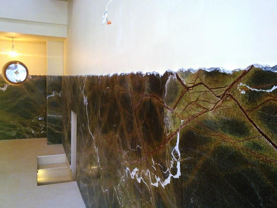 Wnętrze wykończone zielonym kamieniem z brązowymi żyłkami, tworzącym nieregularną krawędź na styku z białą ścianą, widoczny fragment schodów i okrągłe lustro.