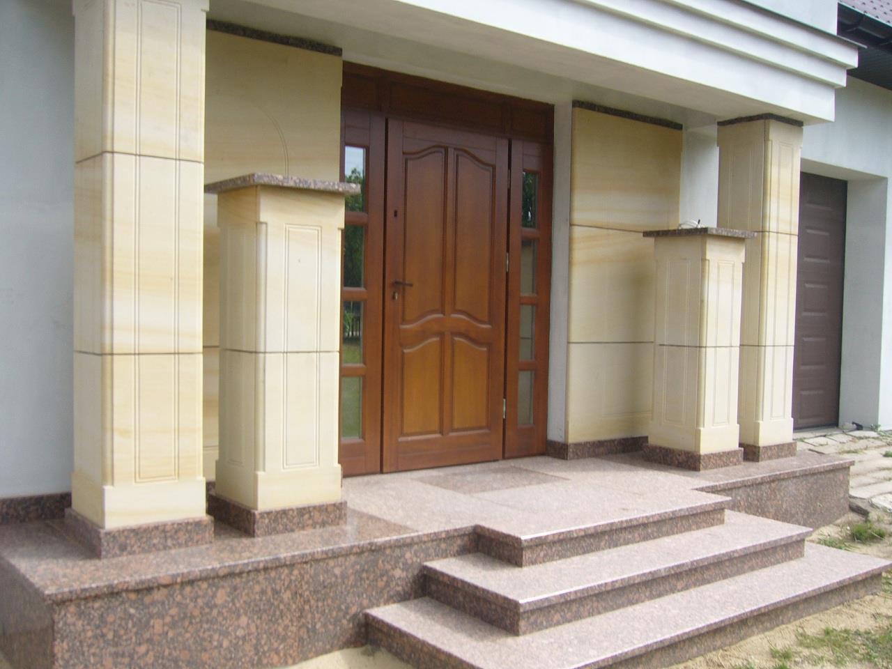 Wejście do domu z drewnianymi drzwiami, kolumnami z piaskowca i schodami z brązowego granitu, widoczne elementy wykończenia elewacji.
