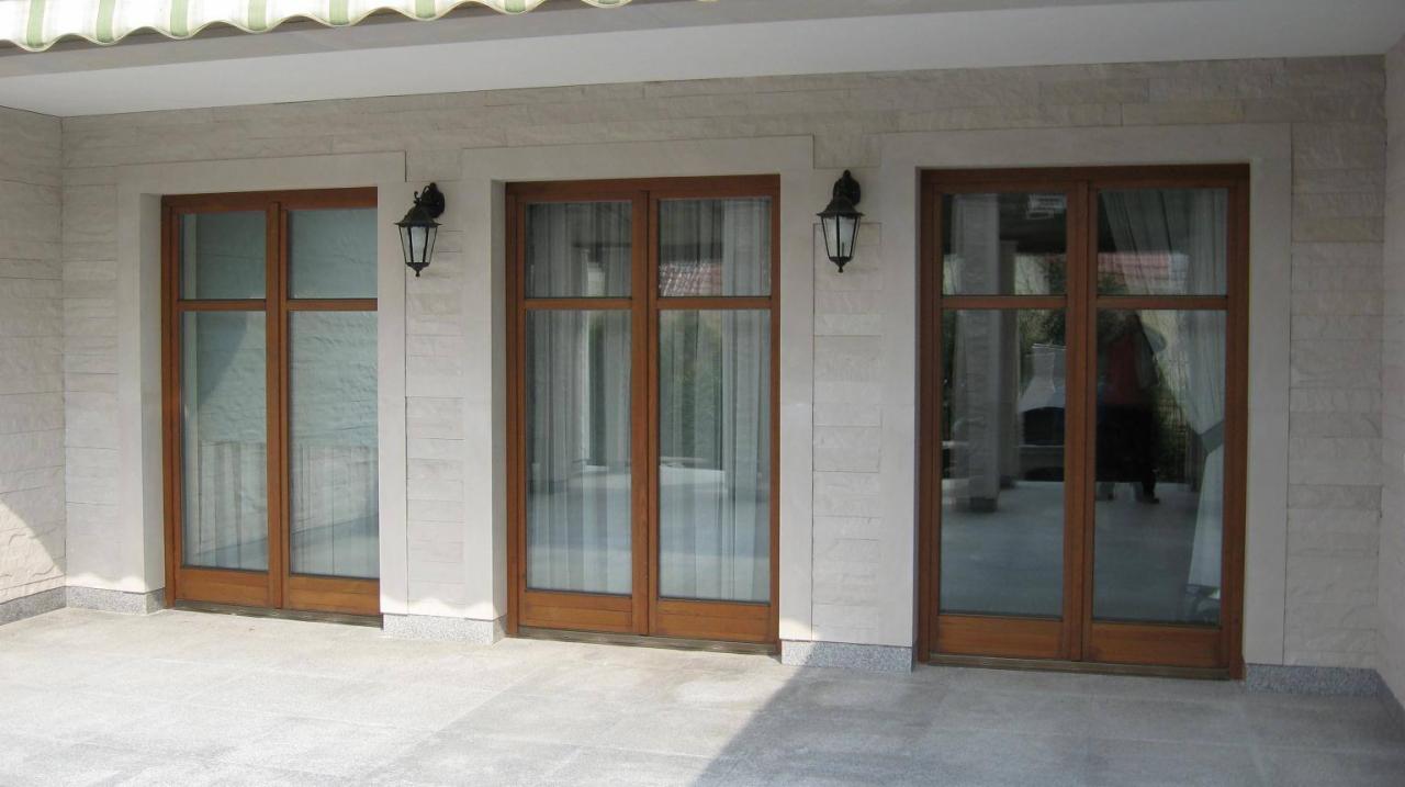 Trzy przeszklone, drewniane drzwi balkonowe w jasnej, kamiennej elewacji z lampami ściennymi, wykonane przez UNIVERSTONE.