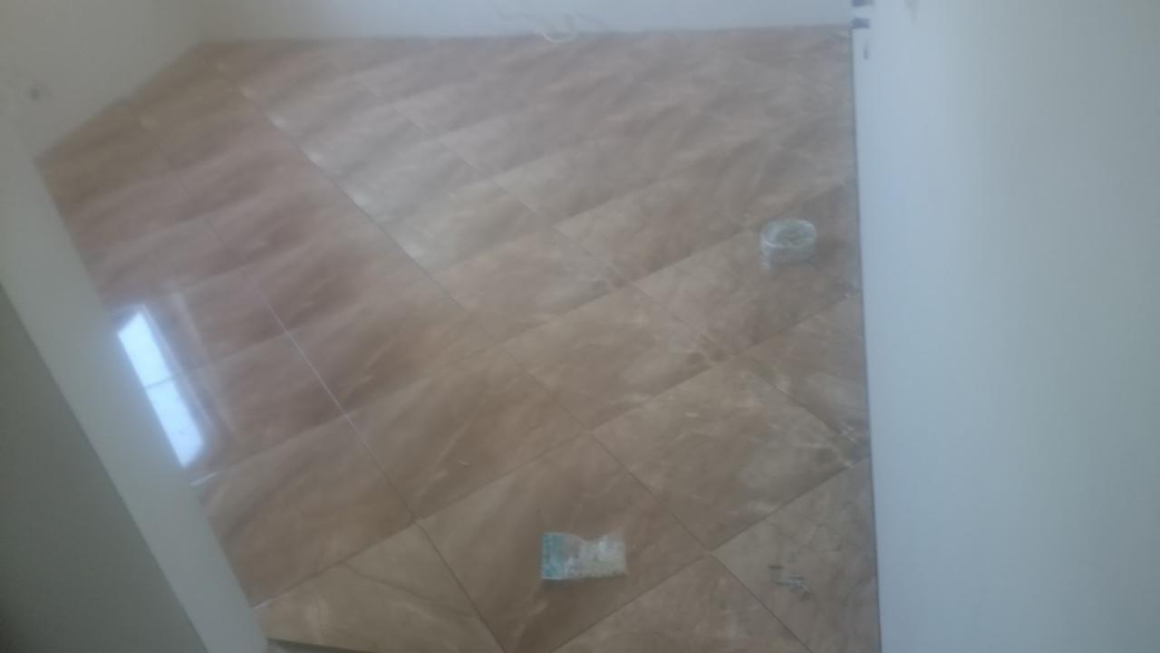 Ułożone, beżowo-brązowe płytki podłogowe w romby, widoczny odblask światła na płytkach, resztki zaprawy i plastikowy pojemnik.