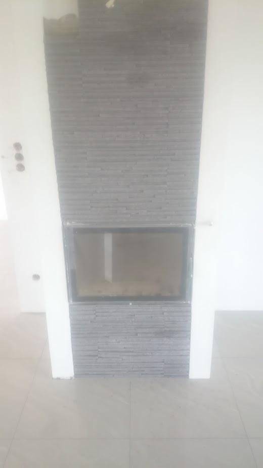 Obudowa kominka wykończona szarą, kamienną okładziną imitującą cegiełki, z widocznym wkładem kominkowym i fragmentem białej ściany po bokach.