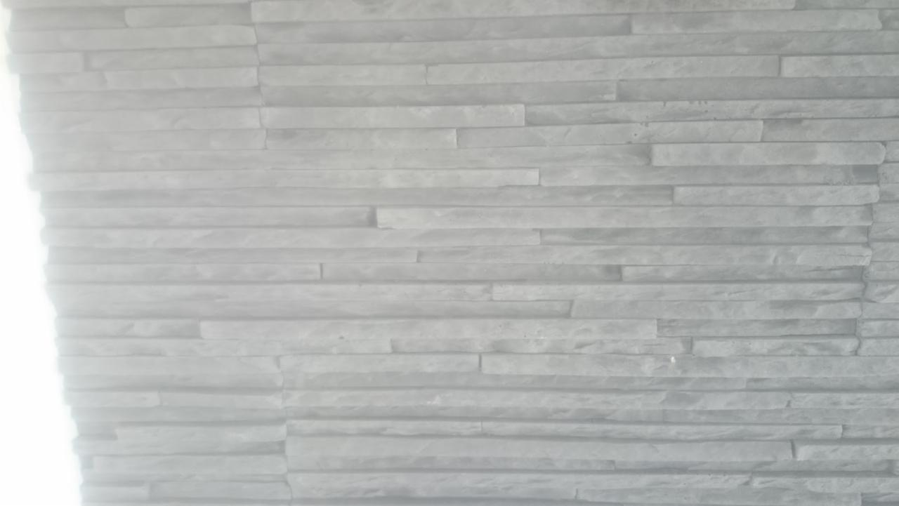 Jasnoszara ściana wyłożona dekoracyjnym kamieniem imitującym cegłę, ułożonym w poziomych pasach z przesunięciem, z jasnym prześwietleniem po lewej stronie.