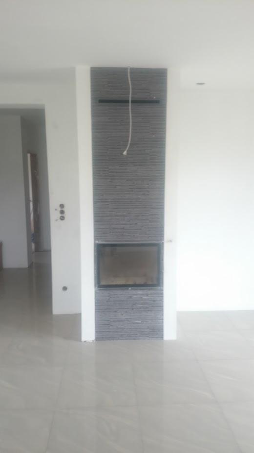 Obudowa kominka wykończona szarą, poziomą glazurą w minimalistycznym wnętrzu, widoczna biała ściana i podłoga z dużych, jasnych płytek.