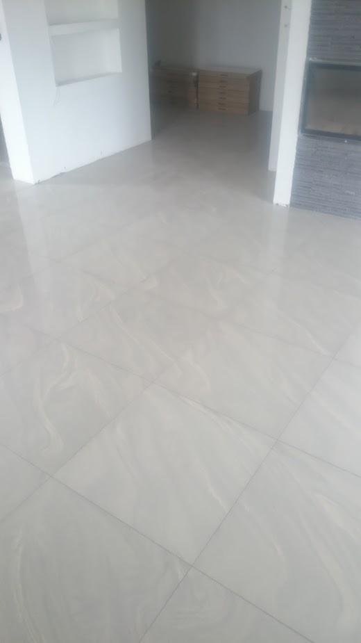 Jasnoszara, błyszcząca podłoga z płytek ceramicznych imitujących marmur, ułożonych w kwadraty, z widocznymi fugami; w tle sterta zapakowanych płytek i fragment kominka obłożonego szarym kamieniem.