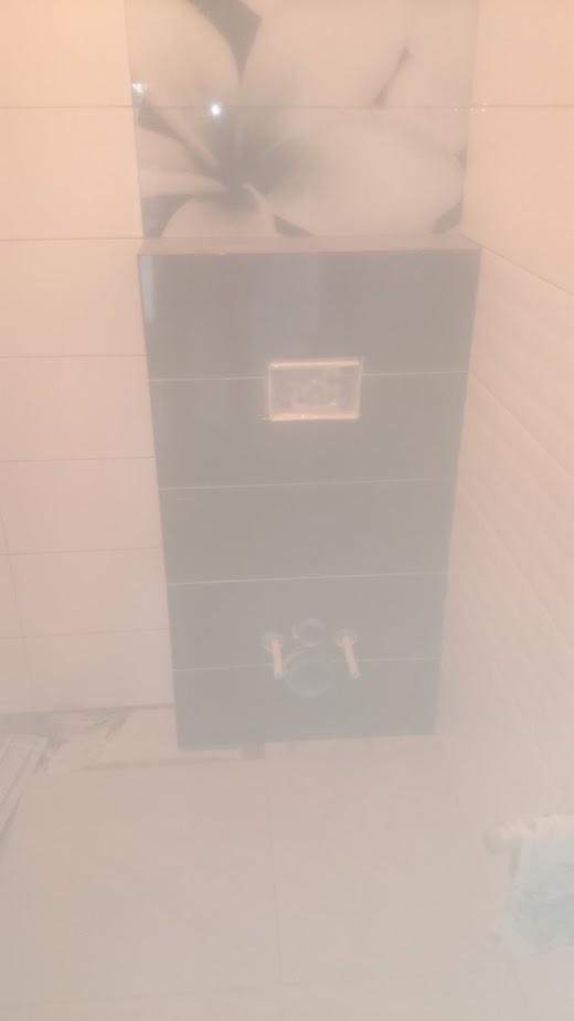 Ciemna obudowa stelaża podtynkowego WC z widocznymi przyłączami hydraulicznymi, na ścianie jasne płytki z motywem kwiatowym.