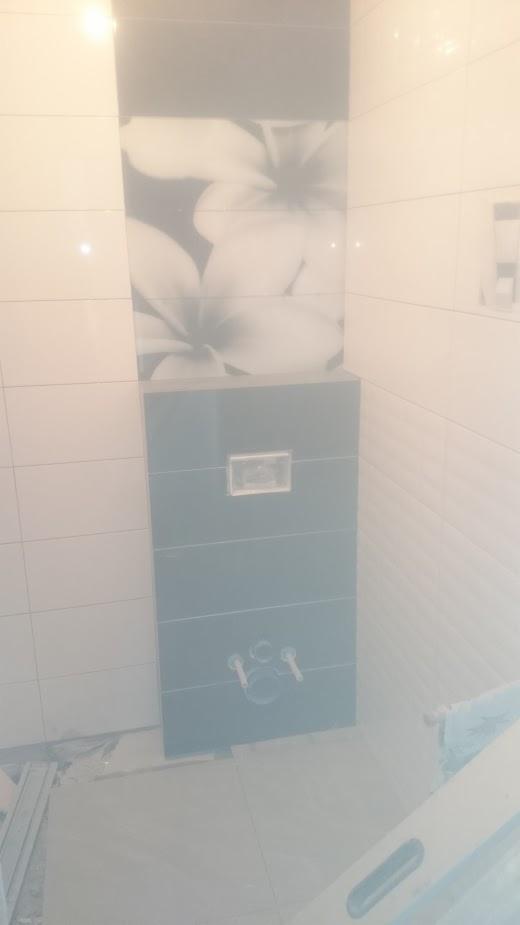 Ściana w łazience z fragmentem pionowego pasa płytek: górna część z motywem kwiatowym, dolna w kolorze szarym z przyciskiem spłuczki i wystającymi rurami. Na podłodze płyty przed położeniem.