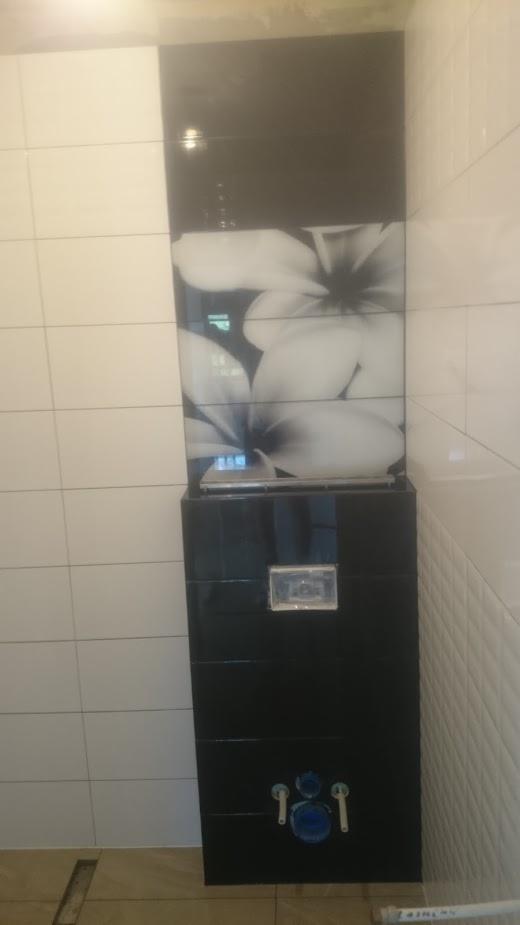 Wykończona łazienka: Białe i czarne płytki ścienne z motywem kwiatowym nad zabudowaną toaletą podtynkową z chromowanymi przyciskami i armaturą.