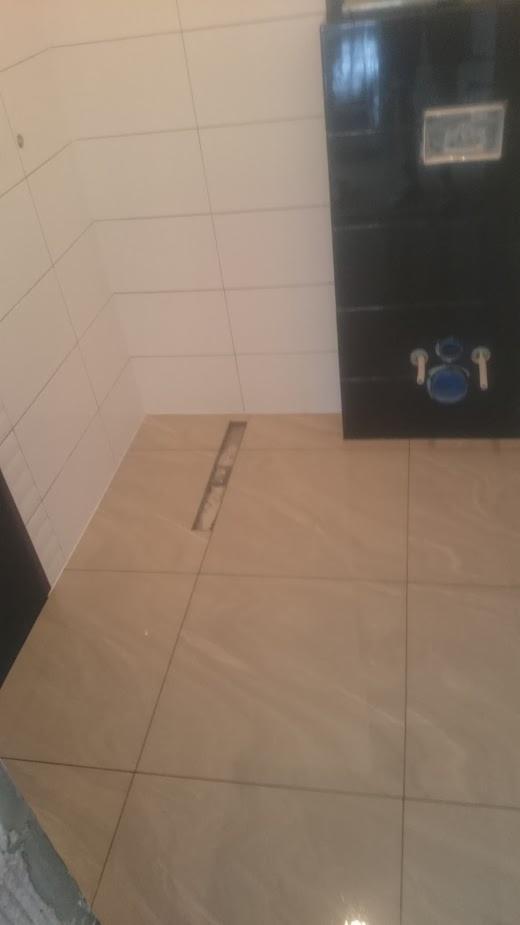 Beżowe płytki podłogowe w łazience z odpływem liniowym w trakcie instalacji, białe płytki ścienne w górnej części, czarna obudowa geberitu.