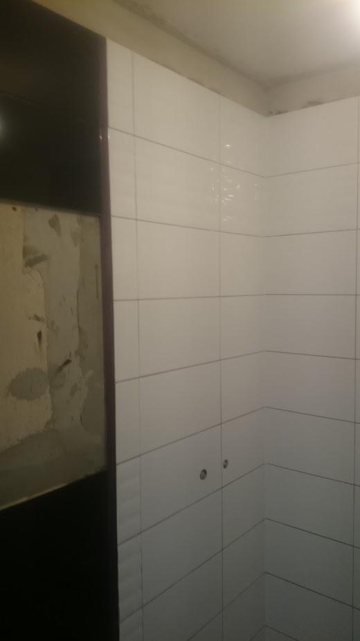 Białe, błyszczące płytki ułożone na ścianie łazienki, widoczne otwory na rury i fragment ściany przed położeniem płytek.