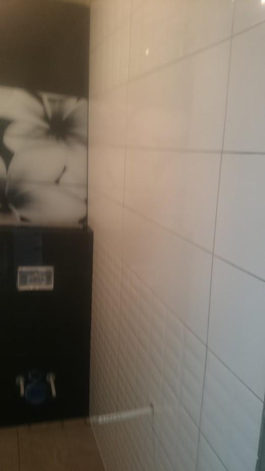 Ściana wyłożona białymi, prostokątnymi płytkami z delikatnym wzorem 3D, widoczny fragment czarnej szafki z grafiką kwiatową i wystającym elementem instalacji wodnej.