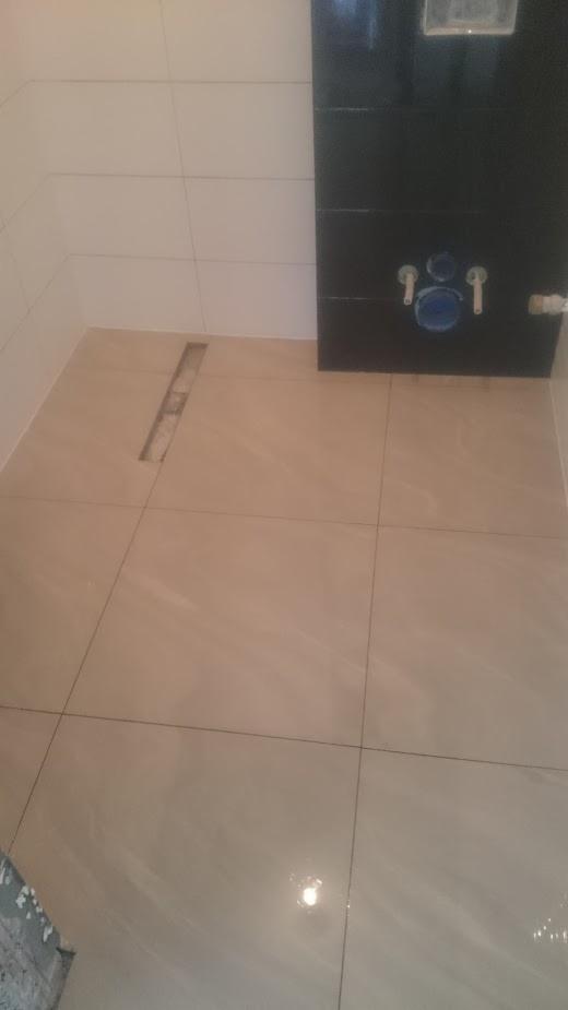 Beżowe płytki podłogowe w łazience z widocznym odpływem liniowym i fragmentem ściany z czarnymi płytkami oraz instalacją podtynkową.