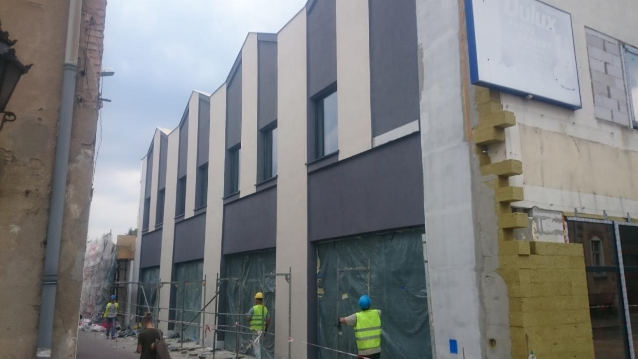 Budynek w trakcie remontu elewacji, widoczne rusztowania, ocieplenie i fragmenty starej i nowej fasady z szaro-białymi pasami. Pracownicy w kamizelkach odblaskowych.