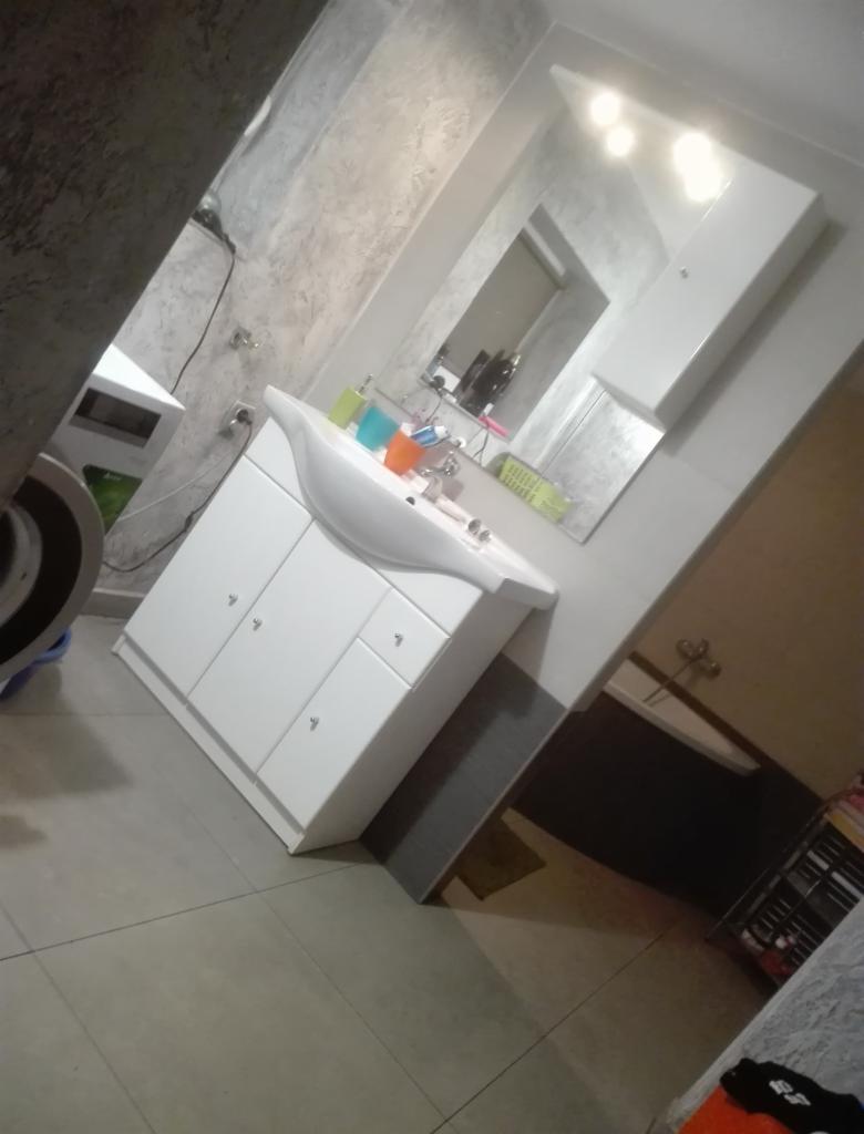 Ukończona łazienka z białą szafką pod umywalką, lustrem z oświetleniem i szarą glazurą na ścianach. Widoczny fragment wanny w tle.