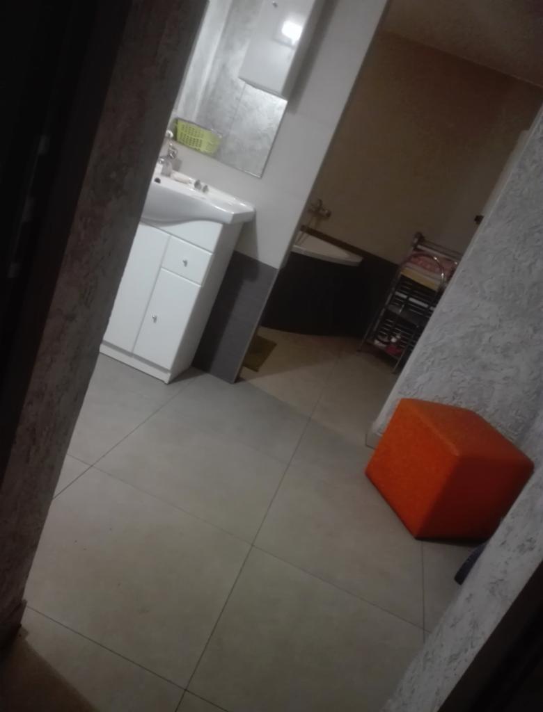 Wykończona łazienka z białymi płytkami podłogowymi, białą szafką pod umywalką, lustrem, półką na kółkach i pomarańczowym pufem w rogu pomieszczenia.