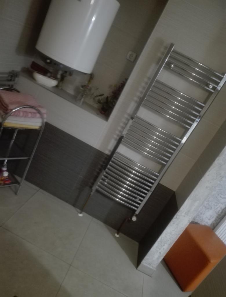 Łazienka po remoncie: ciemnoszare płytki na ścianie, jasne płytki na podłodze, grzejnik drabinkowy, bojler elektryczny, półka z ręcznikami, pomarańczowy puf.