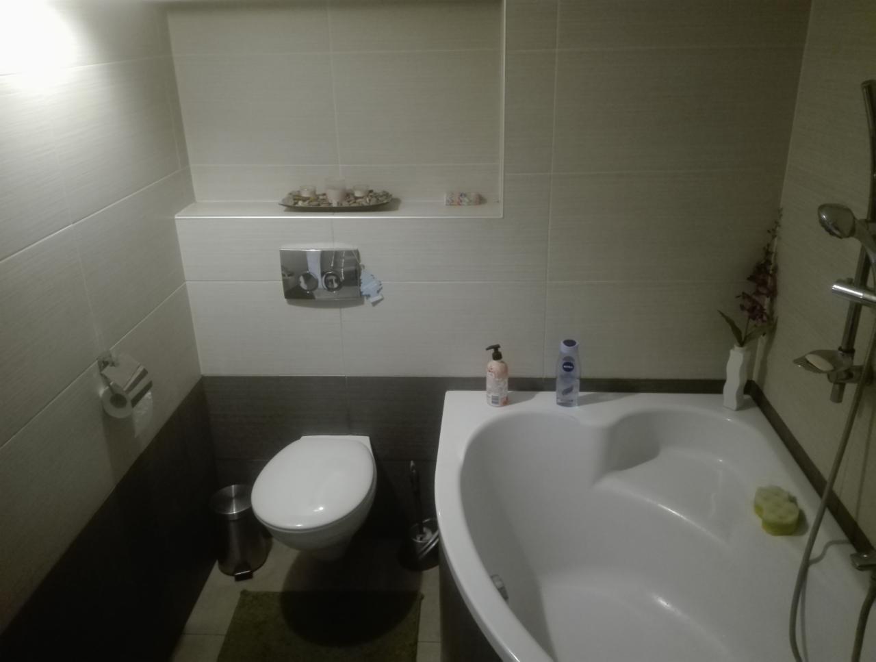 Wykończona łazienka w Krobii z białą armaturą, jasnymi płytkami i ciemniejszymi panelami dolnymi. Na półce nad toaletą dekoracyjne kamienie, a przy wannie kosmetyki i kwiaty.