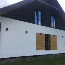 Nowoczesny dom z białą elewacją, czarnym dachem i elementami drewnianymi, oświetlenie zewnętrzne w postaci małych czarnych lamp.