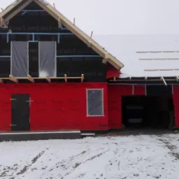 Surowy stan domu szkieletowego w zimowej scenerii, widoczne czerwone płyty izolacyjne Corotop na ścianach, czarna membrana Delta na poddaszu, częściowo ośnieżony dach i zaparkowany samochód.