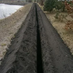 Wykopany rów w ziemi wzdłuż brzegu zbiornika wodnego, przygotowany pod instalację systemu nawadniania.