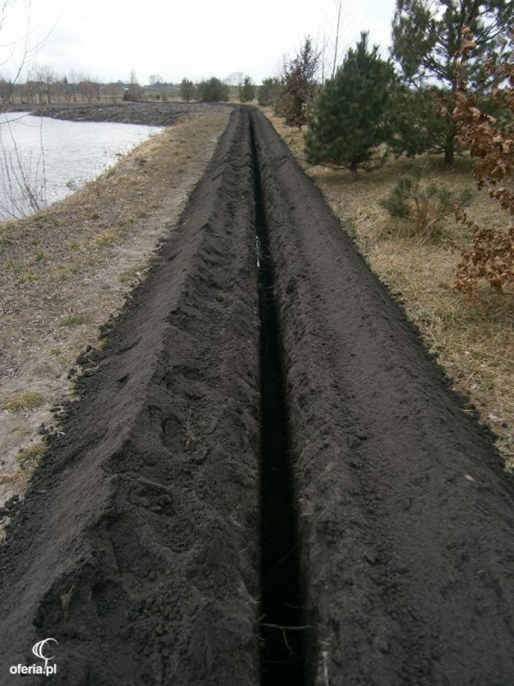 Wykopany rów w ziemi wzdłuż brzegu zbiornika wodnego, przygotowany pod instalację systemu nawadniania.