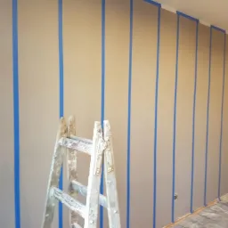 Ściana przygotowana do malowania z użyciem niebieskiej taśmy malarskiej, widoczna drewniana drabina malarska.