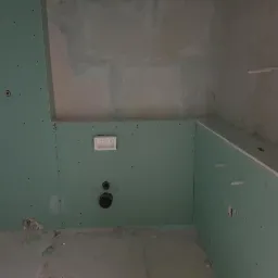 Wnętrze łazienki w trakcie remontu: ściany wyłożone zieloną płytą kartonowo-gipsową, widoczne otwory na instalacje, surowy stan wykończenia.