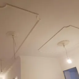 Biały sufit z dekoracyjnymi gładziami w kształcie prostokątów z zaokrąglonymi rogami i rozetami, widoczne kable od lampy wiszącej, wnętrze w trakcie wykończenia.