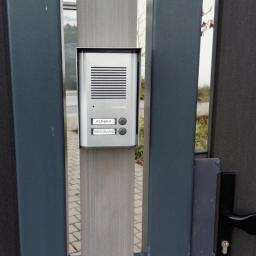 Monser - Szary domofon Commax z przyciskami 'Klinika' i 'Mieszkanie', zamontowany na metalowym słupku ogrodzenia.