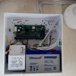 Otwarta obudowa centrali alarmowej z widoczną płytą główną, transformatorem, akumulatorem oraz okablowaniem; obok zamontowany czujnik alarmowy.