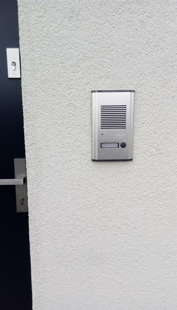 Srebrny domofon z przyciskiem i głośnikiem zamontowany na białej, szorstkiej ścianie obok fragmentu ciemnych drzwi z metalową klamką.