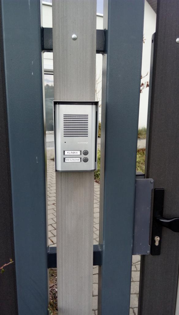 Szary domofon Commax z przyciskami 'Klinika' i 'Mieszkanie', zamontowany na metalowym słupku ogrodzenia.