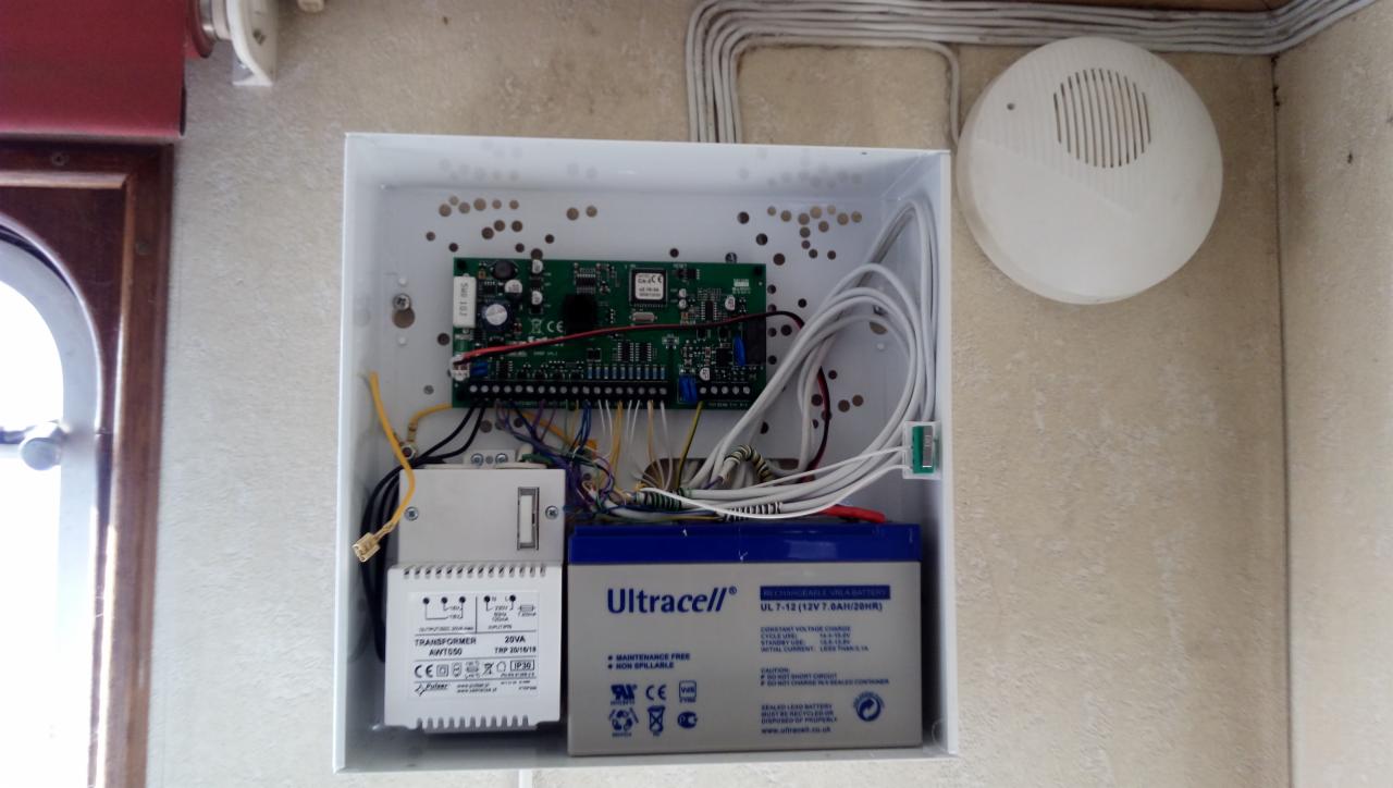 Otwarta obudowa centrali alarmowej z widoczną płytą główną, transformatorem, akumulatorem oraz okablowaniem; obok zamontowany czujnik alarmowy.