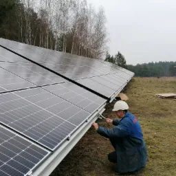 Instalator w kasku dokręca śruby mocujące panele słoneczne do konstrukcji na tle lasu w pochmurny dzień.