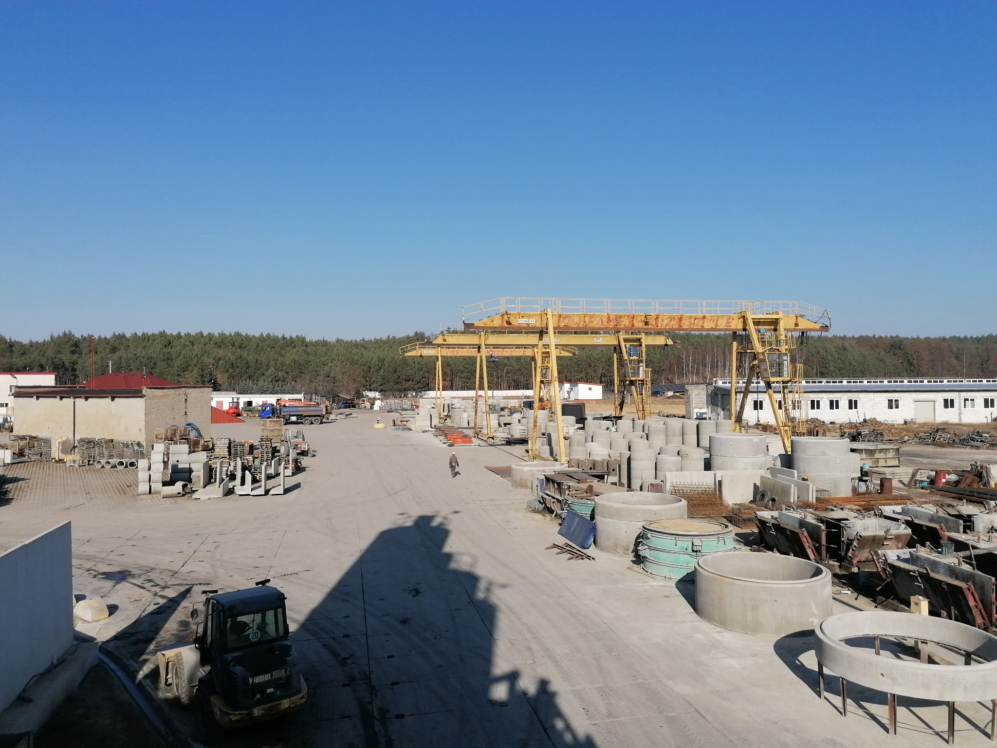 Plac składowy z betoniarnią, widok z góry na różnorodne prefabrykaty betonowe, w tym kręgi betonowe, płyty i inne elementy konstrukcyjne; w tle las i błękitne niebo, na pierwszym planie cień wózka...