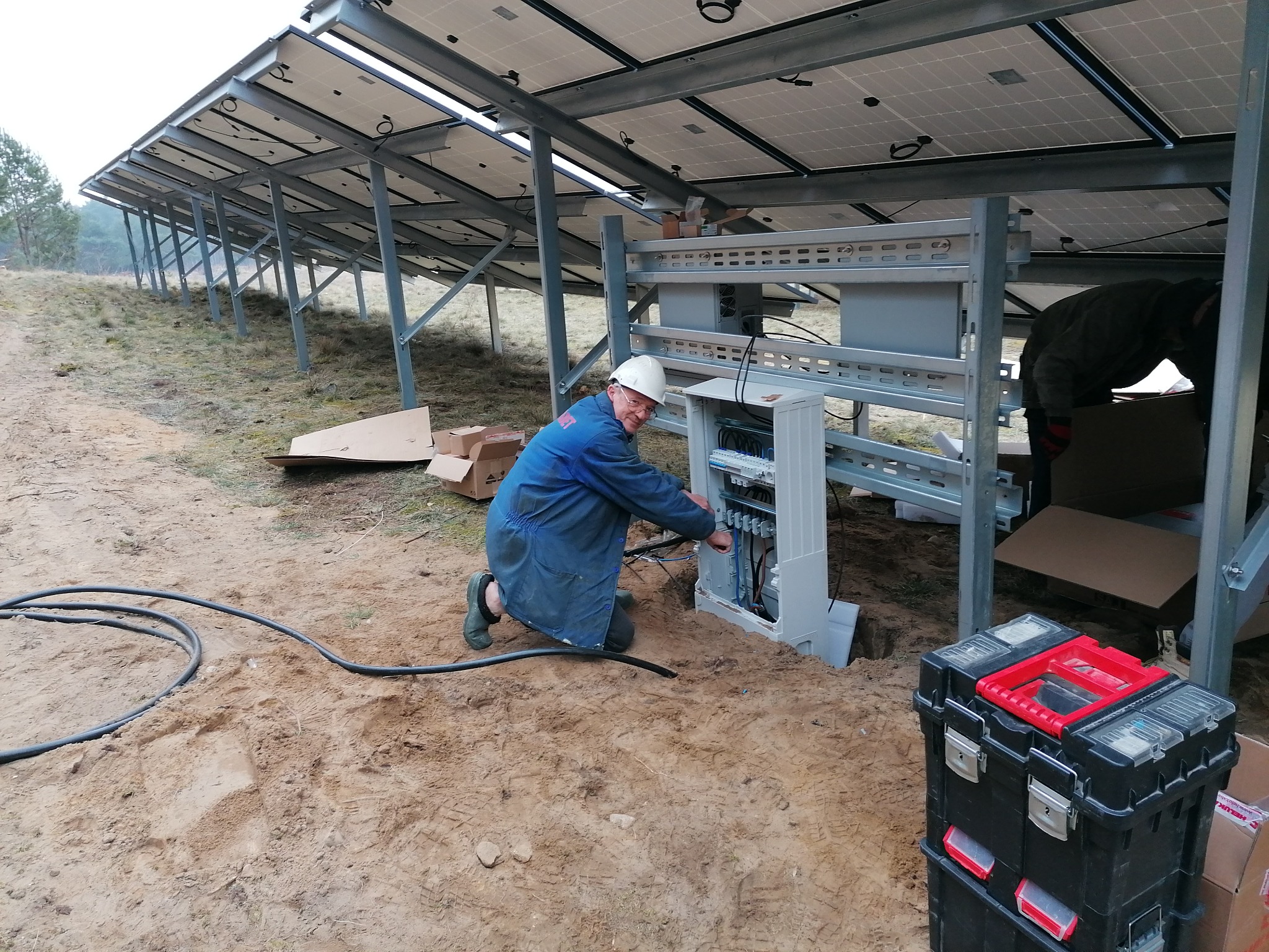 Instalacja paneli słonecznych: elektryk w kasku podłącza przewody w szafie sterowniczej, widoczne panele na metalowej konstrukcji i skrzynka z narzędziami na piaszczystym podłożu.