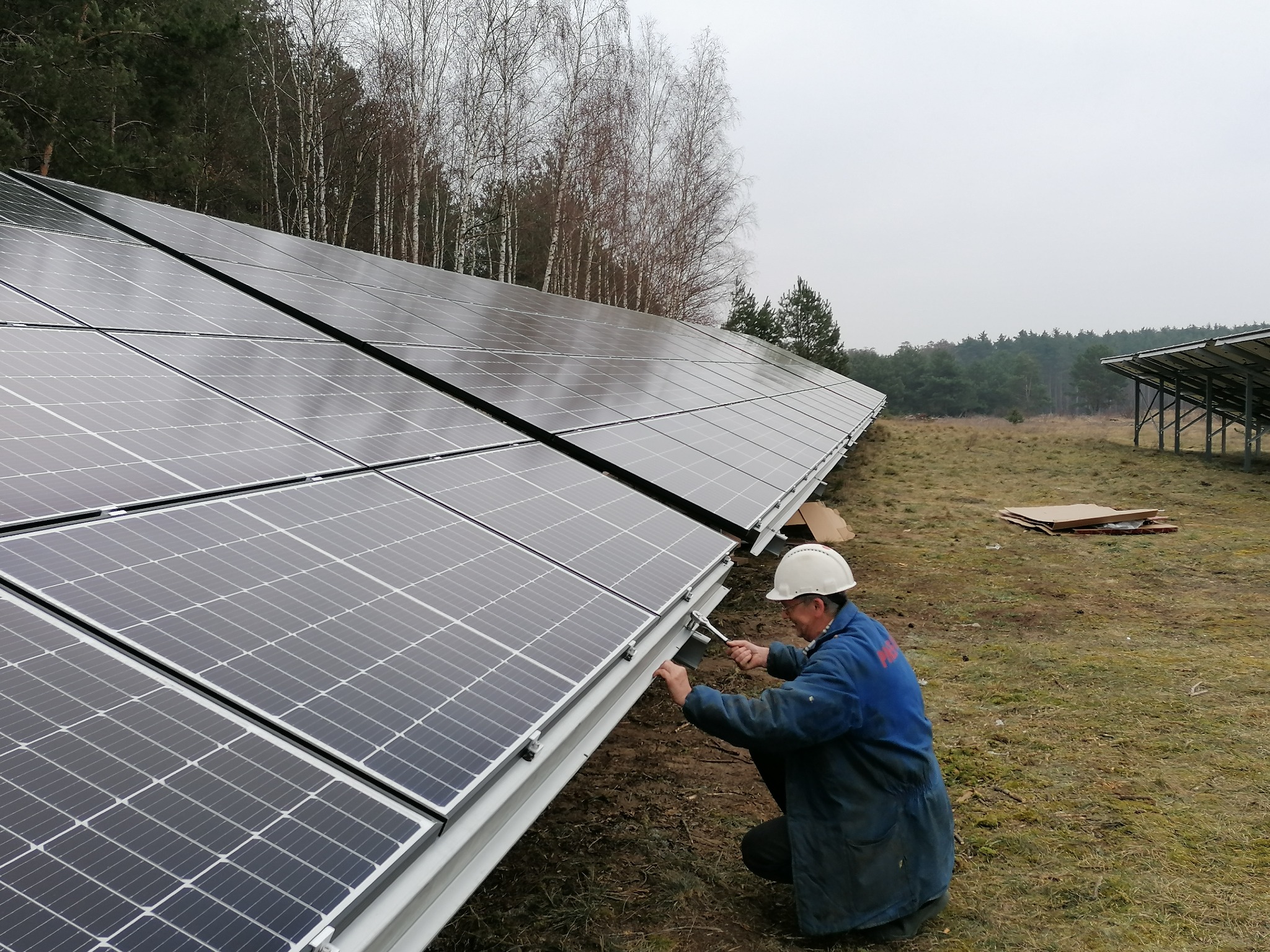 Instalator w kasku dokręca śruby mocujące panele słoneczne do konstrukcji na tle lasu w pochmurny dzień.